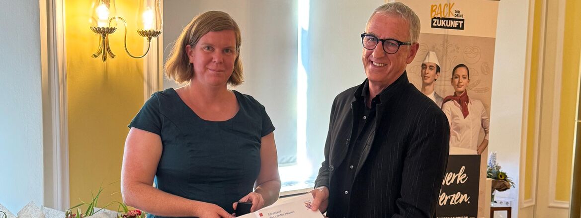 Hans-Hermann Schröer erhält den Ehrenbrief des Landes Hessen aus den Händen von Stadträtin Frau Löbcke. Die Verleihung findet in feierlichem Rahmen mit Blumen und Roll-up im Hintergrund statt.